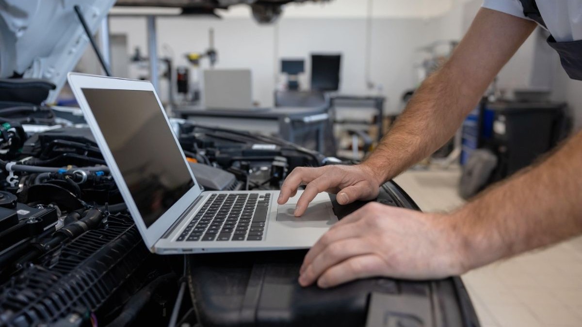 Un mecanic folosește un laptop de culoare gri, plasat pe motorul unei mașini cu capota deschisă, pentru a diagnostica problema, într-un service auto