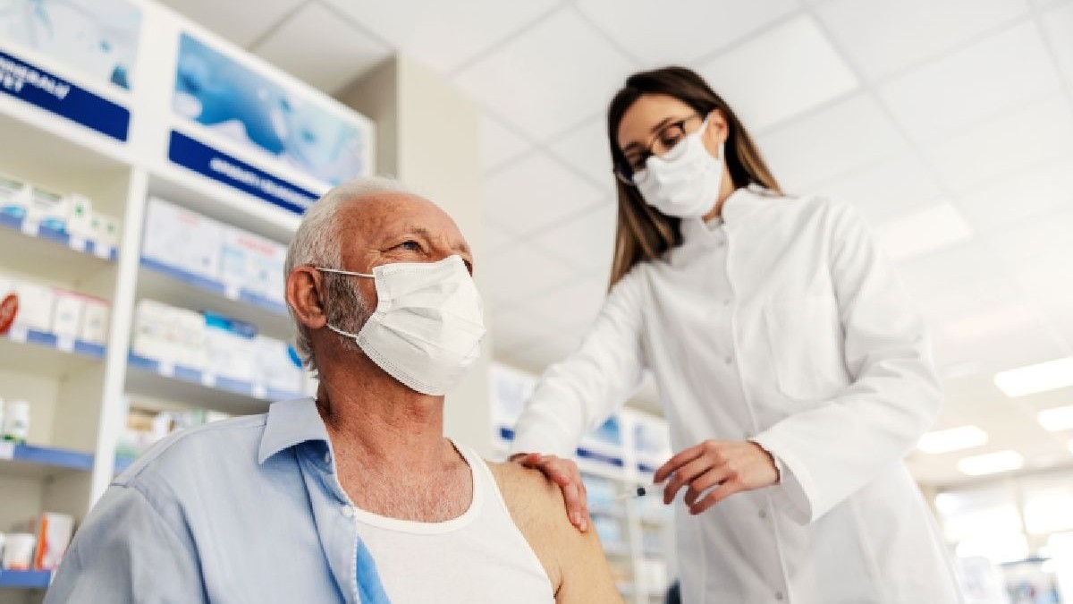 Un barbat cu masca la gura sta pe scaun intr-o farmacie, cu bratul dezgolit si o femeie imbracata in halat alb si cu masca la gura ii face un vaccin in brat. Vaccinare antigripala in farmacii. Foto: Shutterstock