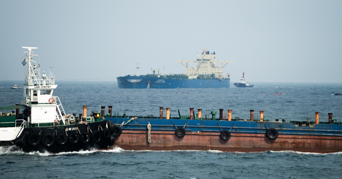 Imaginea prezintă în plan apropiat o barjă lungă flancată de un remorcher de manevră, iar în fundal un cargobot de mari dimensiuni care navighează în larg.