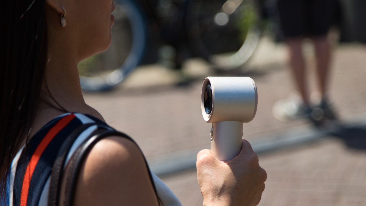 Profilul unei femei care ține în mână un mini-ventilator portabil de culoare deschisă, în timp ce se află în aer liber într-o zonă pietonală estompată în fundal.