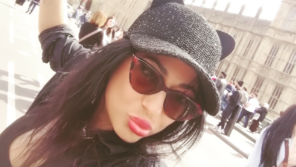 Imaginea este un selfie realizat de o femeie cu părul lung și negru, care poartă o șapcă neagră cu urechi de pisică și ochelari de soare cat-eye cu lentile roșiatice. Pe fundal, se poate distinge o clădire istorică mare, probabil Palatul Westminster din Londra, sugerând că fotografia a fost făcută într-o zonă turistică aglomerată.