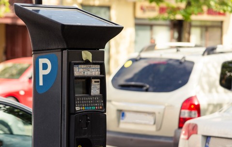 Prim-plan cu un aparat electronic de plată a parcării, alimentat cu panou solar, amplasat pe marginea unei străzi cu mașini parcate.