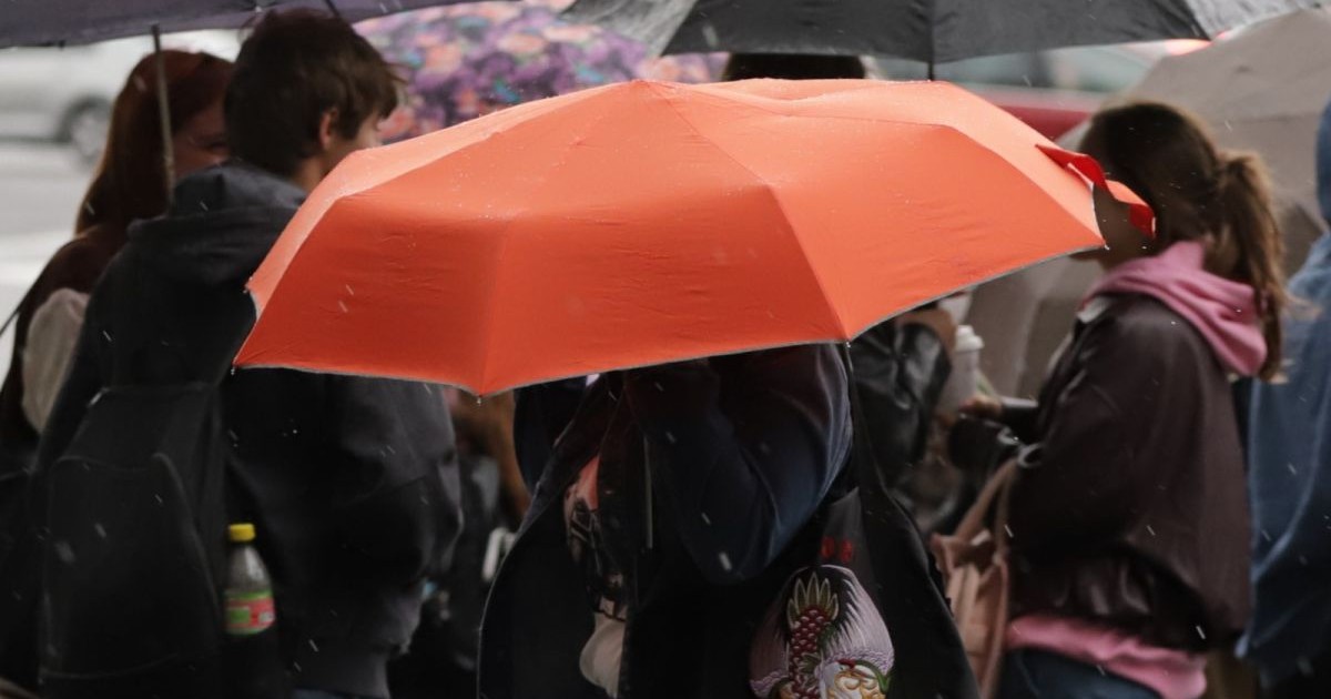 Mai mulți tineri fotografiați cu spatele sau din profil stau sub umbrele, iar în centru se vede o umbrelă mare roșie-portocalie, într-o zi ploioasă rece, București, 29 septembrie 2025