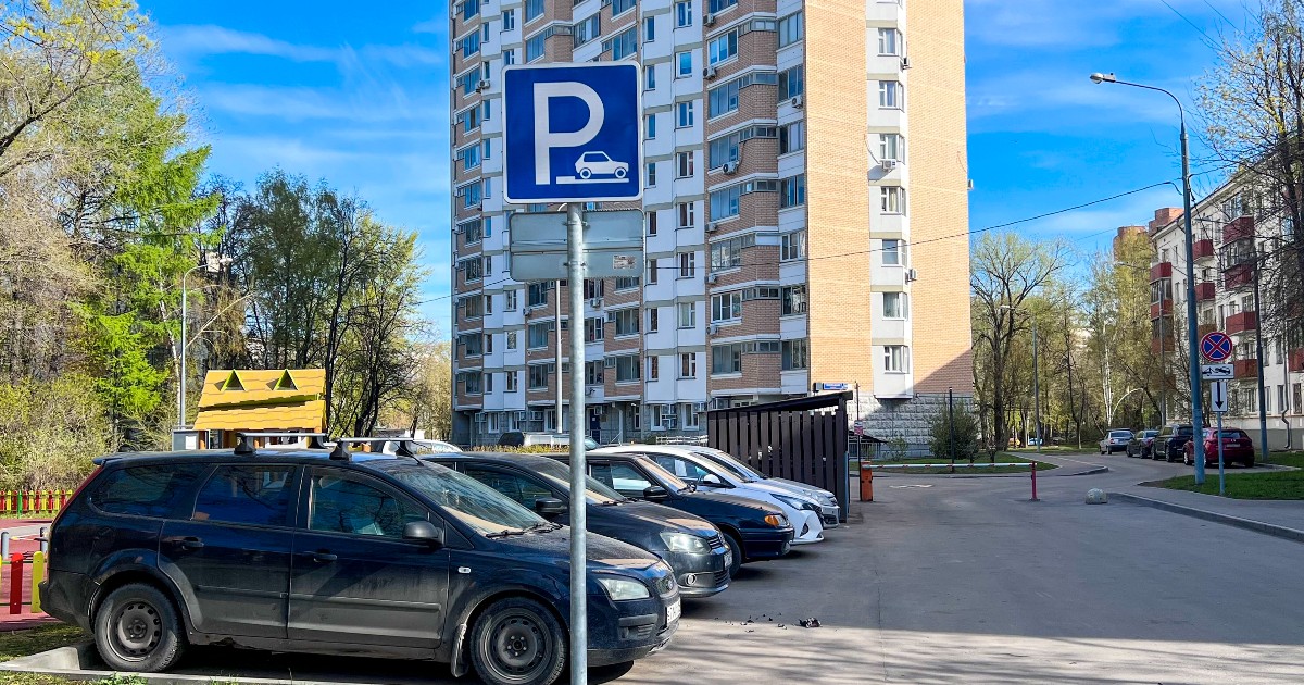 Imaginea prezintă un indicator albastru de parcare amplasat în fața unui rând de mașini staționate pe trotuar, lângă un bloc înalt de locuințe.