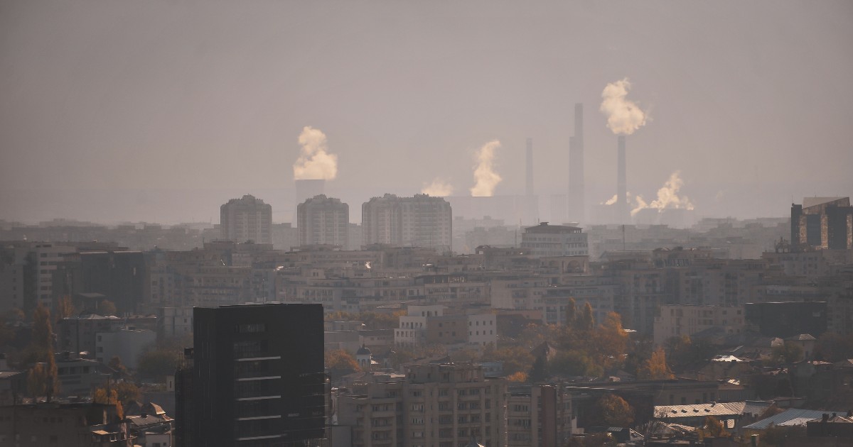 Imaginea prezintă o panoramă urbană densă, dominată de un strat gros de ceață sau smog, care conferă scenei o nuanță cenușie și estompată. În fundal se înalță coșuri de fum industriale înalte, din care iese vizibil abur sau fum, sugerând o sursă majoră de poluare deasupra zonei rezidențiale.