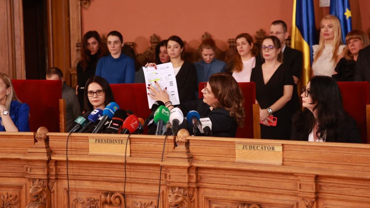 Un grup de oameni în picioare, în spatele a două femei care stau la un prezidiu și una dintre ele arată câteva hârtii pe care scrie ceva