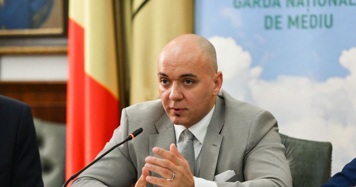 Andrei Corlan, șeful Gărzii Naționale de Mediu, îmbrăcat cu un costum de culoare gri, cu o cămașă albă și cravată gri, cu steagul României pe fundal și un microfon în față, în timp ce stă la o masă, așezat pe un scaun
