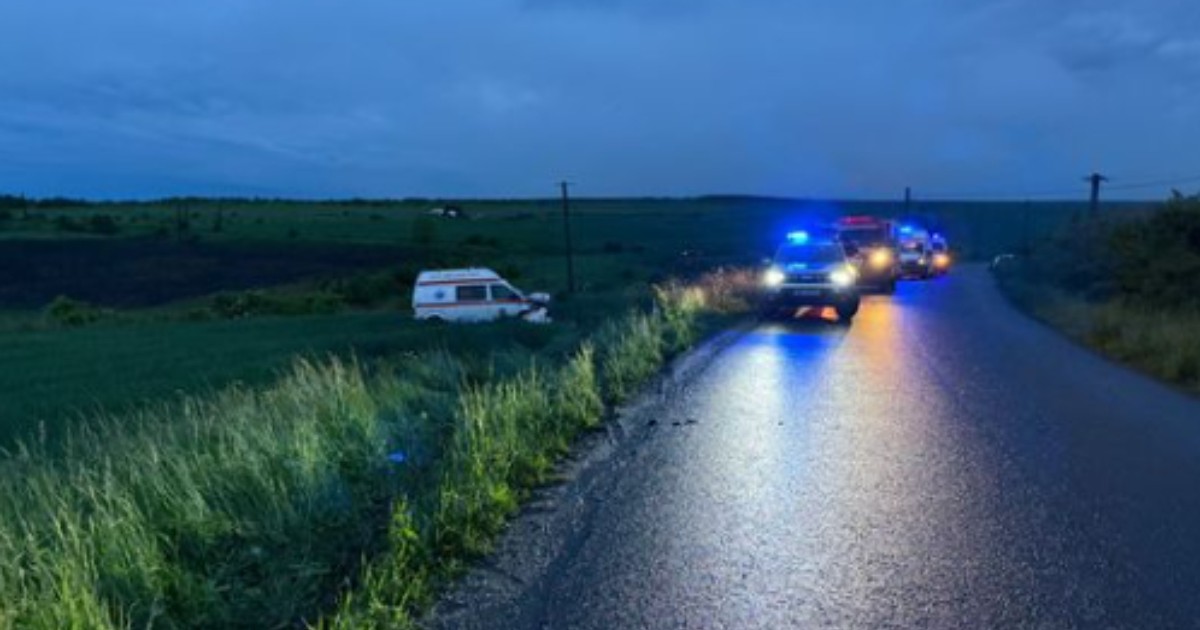 În partea stângă, într-un câmp, se observă o ambulanță ieșită complet de pe carosabil.