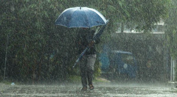 Imaginea surprinde o persoană care înaintează cu greu printr-o ploaie torențială pe stradă, protejându-se cu o umbrelă albastră sub un cer întunecat.
