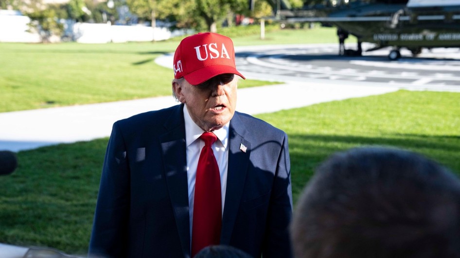 Donald Trump purtând un costum închis la culoare, cravată roșie și o șapcă roșie pe care scrie „USA”, vorbind în aer liber în fața jurnaliștilor, cu un elicopter în fundal.