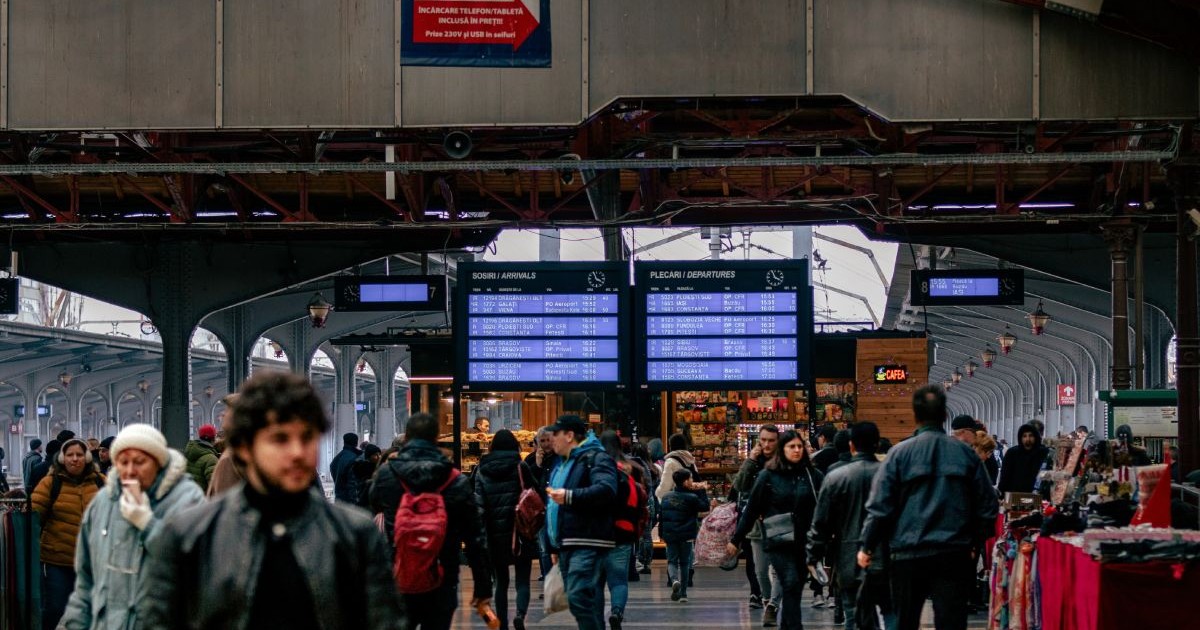 Imagine creativă cu Gara de Nord din București, în prim plan, în blur, mai mulți călători, iar pe fundal panourile de informare cu plecări și sosiri ale trenurilor