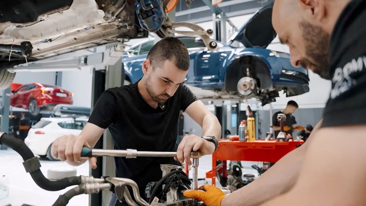 Imaginea prezintă doi mecanici auto lucrând atent la dezasamblarea sau repararea unei componente de motor într-un service auto.