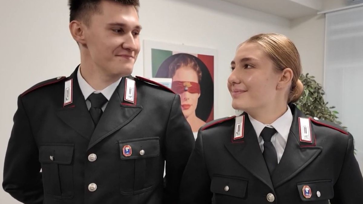 Un tânăr și o tânără îmbrăcați în uniforme negre de poliție, cu epoleți și insigne roșii, zâmbind unul către celălalt