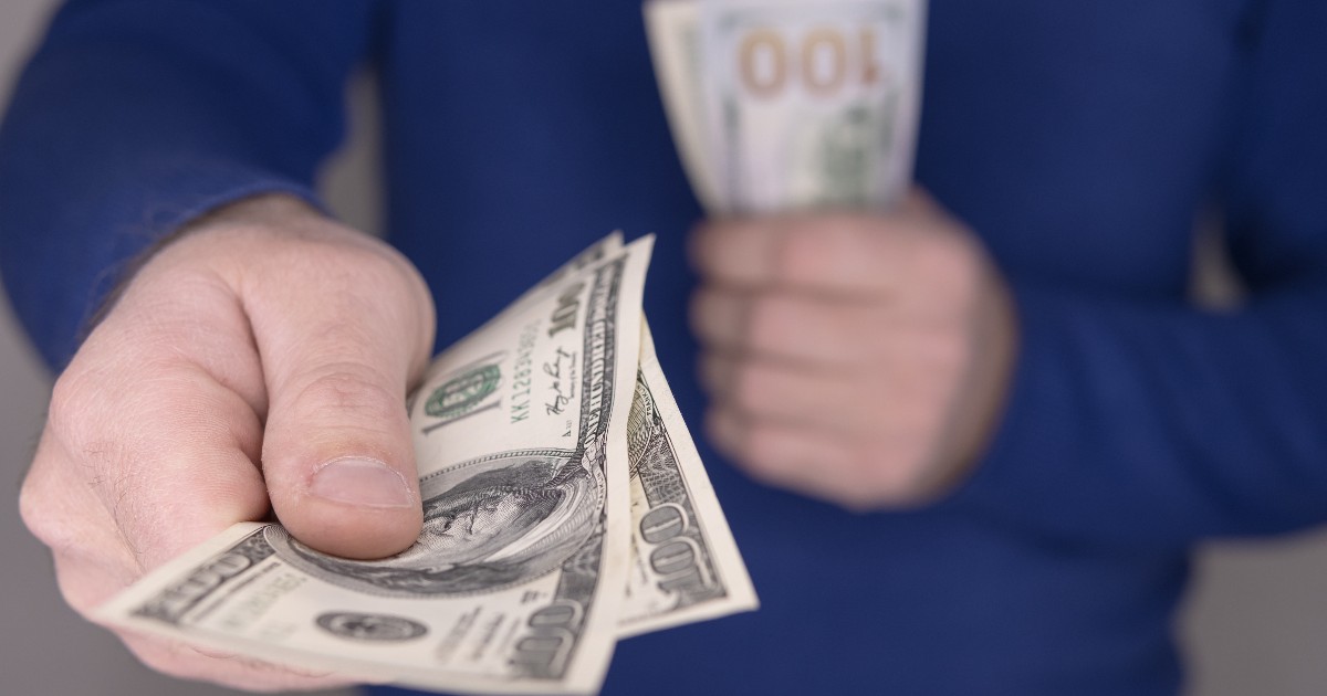 Imaginea surprinde o persoană care întinde câteva bancnote de 100 de dolari, în timp ce în cealaltă mână ține un teanc mai mare de bani. Scena sugerează un gest de plată, împrumut sau oferire de bani.