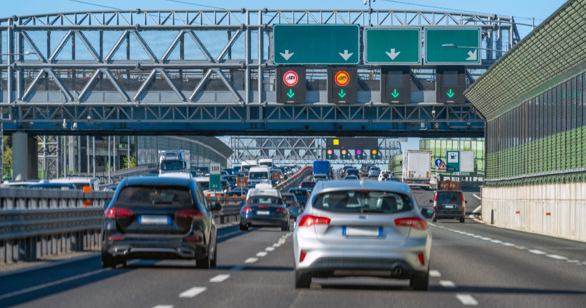 Imaginea prezintă un trafic aglomerat de autoturisme pe o autostradă, trecând pe sub o structură metalică masivă dotată cu panouri electronice și indicatoare rutiere.