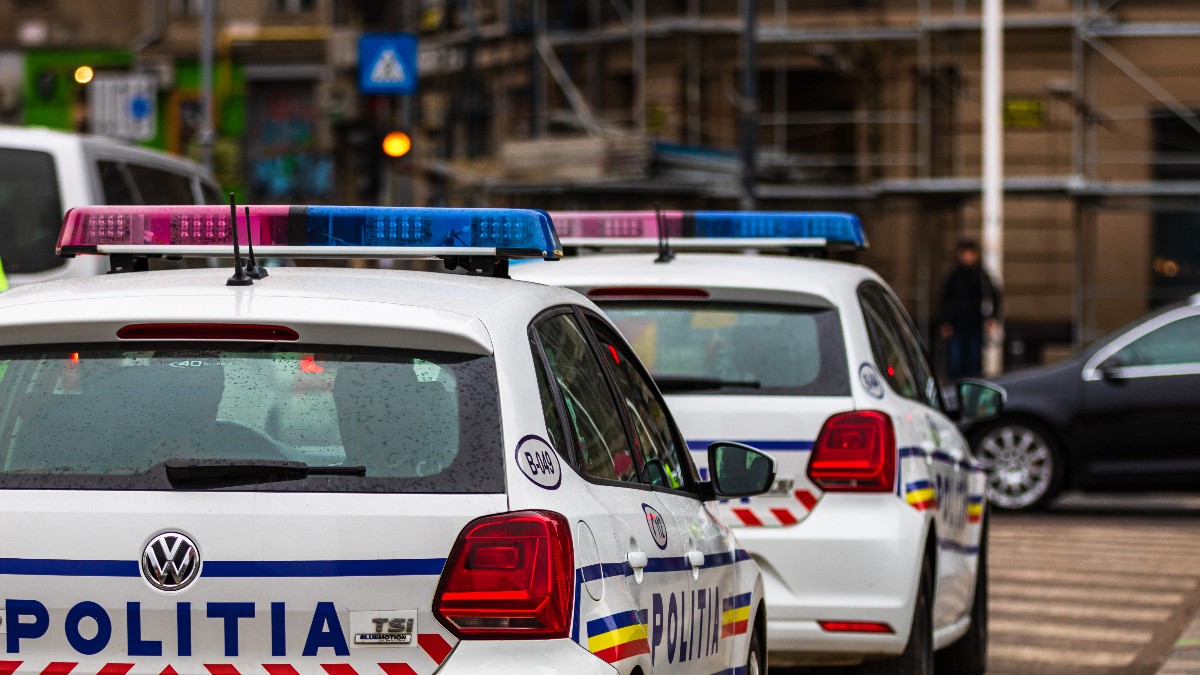 Două mașini de poliție albe, care par a fi modele Volkswagen, sunt aliniate într-un plan apropiat, scoțând în evidență inscripțiile mari "POLIȚIA" cu litere albastre și sistemul de avertizare luminos roșu-albastru de pe plafon. Mașinile au benzi decorative cu culorile drapelului național (roșu, galben, albastru), iar în fundalul urban, estompat, se văd alte vehicule și clădiri în construcție.