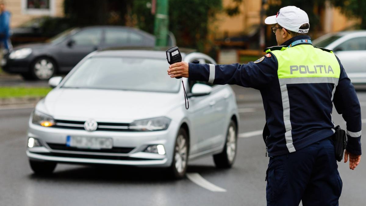 Un polițist rutier oprește un o mașină de culoare argintie, marca Volkswagen, în trafic, Polițistul se afla în fața mașinii, cu mâna stângă ridicată.
