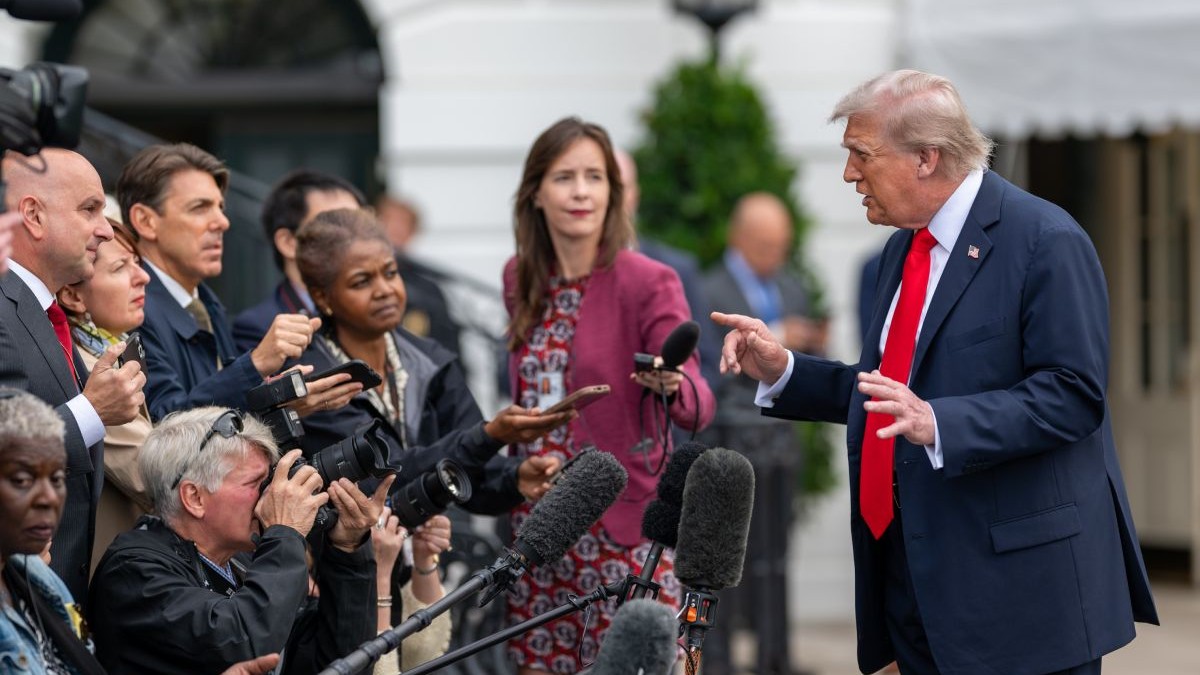 Donald Trump, in dreapta, in costum negru, camasa alba si cravata rosie, sta cu palmele ridicate la nivelul pieptului, fata in fata cu mai multi jurnalisti cu microfoane intinse