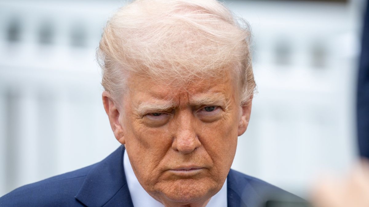 Donald Trump, în prim plan, cu părul alb, și îmbrăcat cu un sacou albastru și o cămașă albă pe un fundal blurat