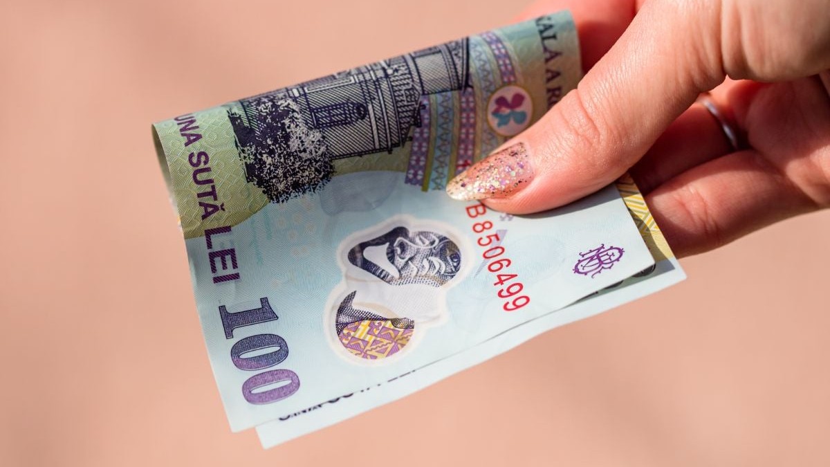 O bancnotă de 100 de lei, împăturită în două, ținută în mână de o femeie, pe fundal roz
