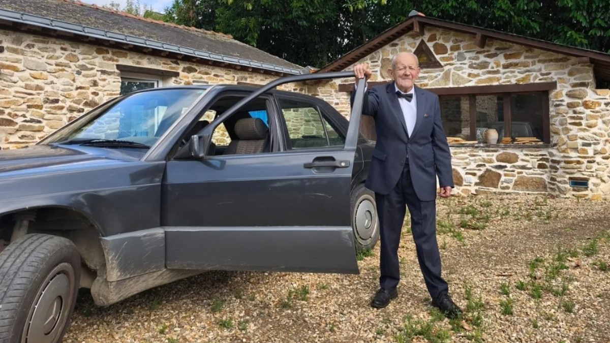Un bărbat în vârstă, îmbrăcat elegant în costum cu papion, stând lângă portiera deschisă a unei mașini, în fața unei case din piatră.