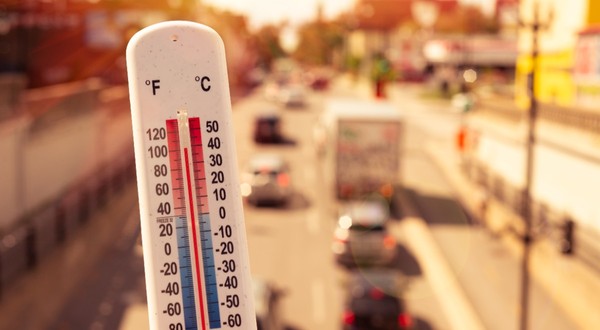 Prim-plan cu un termometru de exterior care indică o temperatură ridicată, pe fundalul unui drum urban însorit și aglomerat cu vehicule.