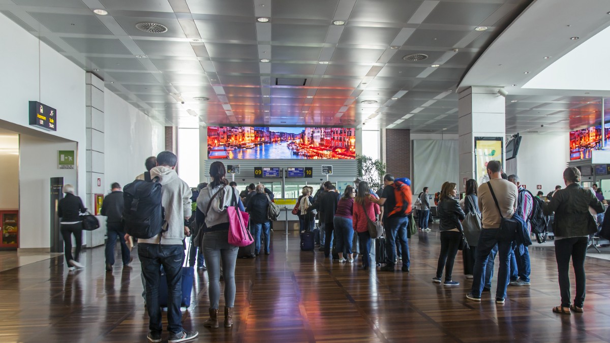 Imaginea prezintă un aeroport aglomerat, cu pasageri care stau la coadă în zona de îmbarcare, ținând bagaje și rucsacuri. Pe fundal se observă ecrane mari cu imagini turistice, iar atmosfera sugerează flux intens de călători.
