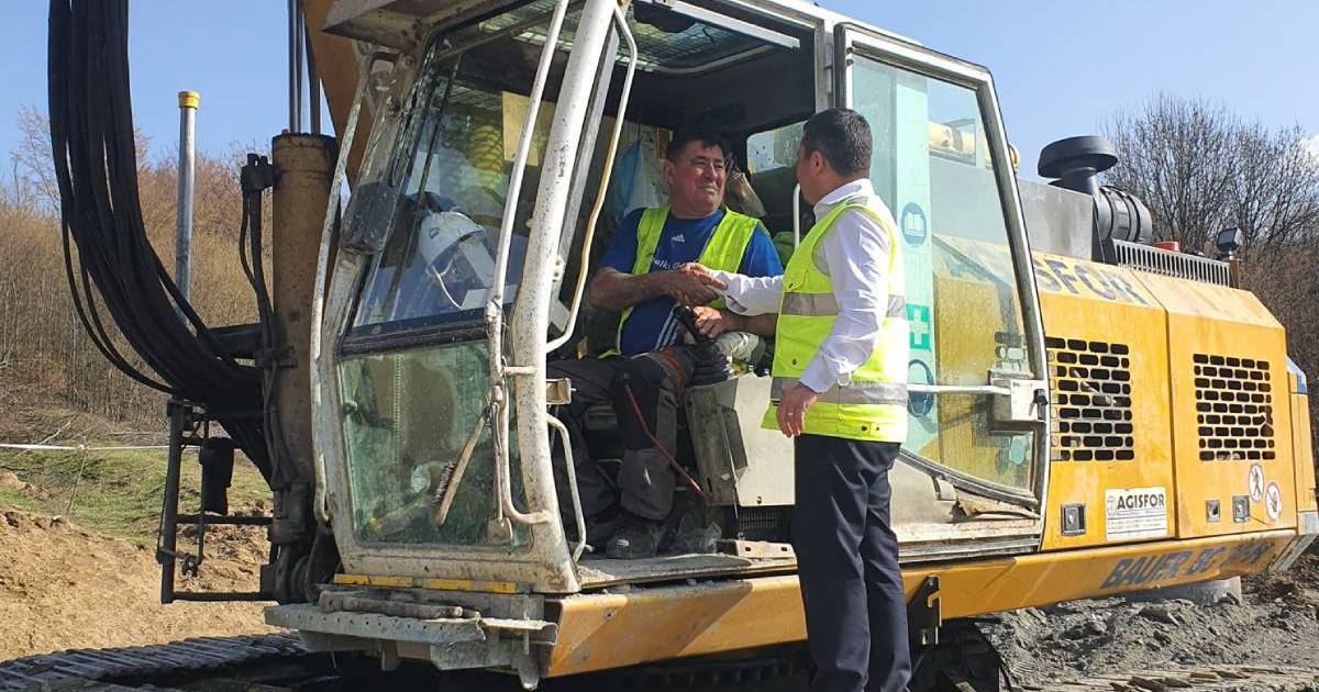 șantier de construcție pe autostrada Sibiu-Pitești, unde un muncitor aflat într-un excavator dă mâna cu un bărbat îmbrăcat în vestă reflectorizantă și cămașă albă