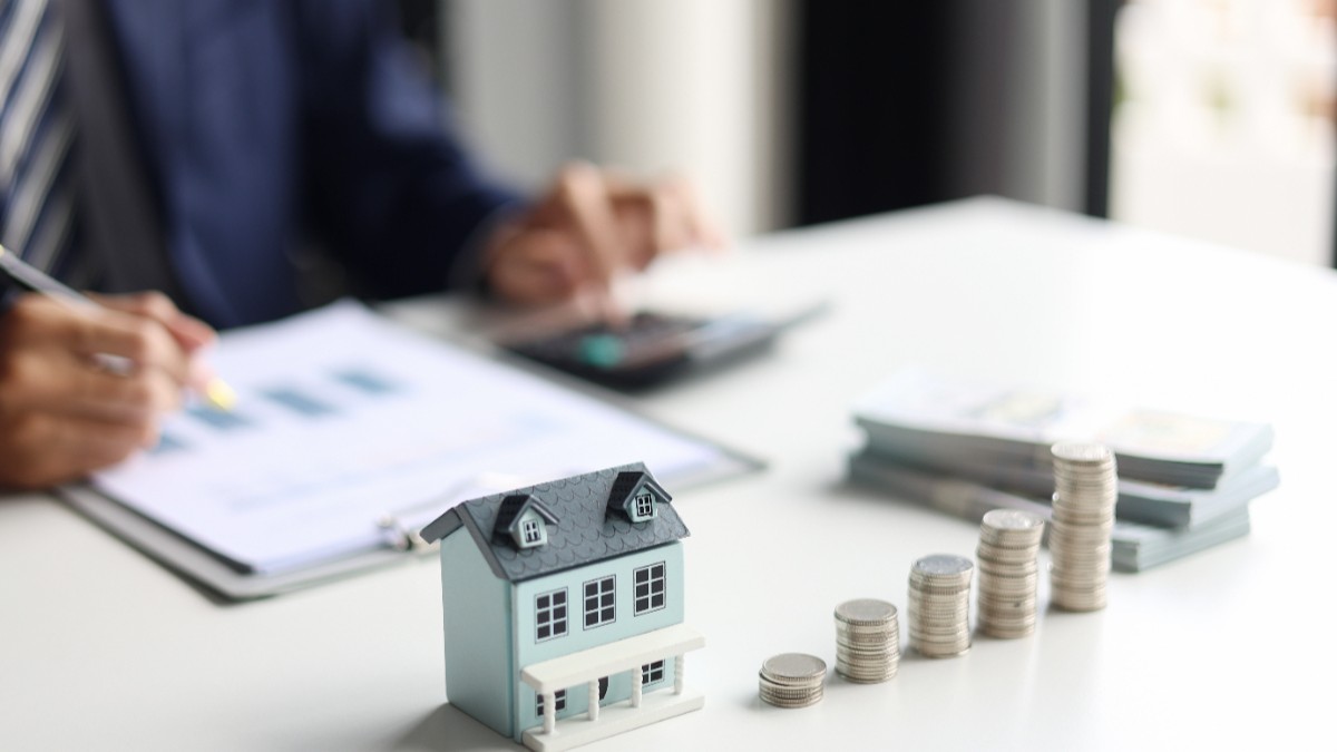 Un agent imobiliar stă la birou cu hârtii în față, are pe masă macheta unei case și mai multe fișicuri de monede așezate unul lângă altul