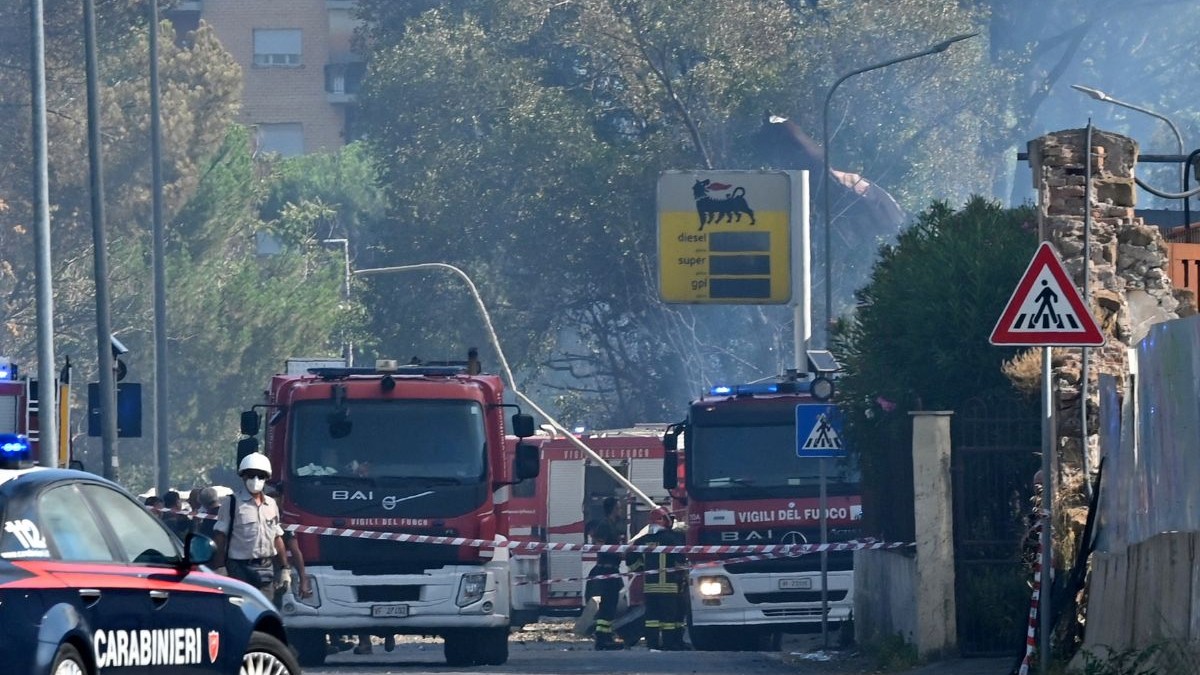 Două autospeciale de stingere a focului sunt staționate în spatele unei benzi colorate în dungi roșii și albe care delimitează zona de pericol. în partea dreaptă a fotografiei se poate observa semnul luminos al unei benzinării ENI, care înfățișează un câine negru pe un fundal galben. în dreapta imaginii se află o mașină neagră de poliție, cu textul „CARABINIERI” în font gros și alb, pe portierele mașinii
