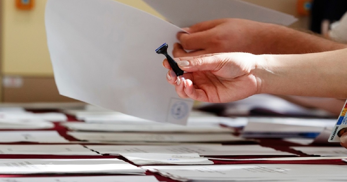 Alegeri prezidentiale 2025, turul 2- Pe un birou se afla mai multe buletine de vot, iar o femeie întinde o stampilă pentru vot unui alegător