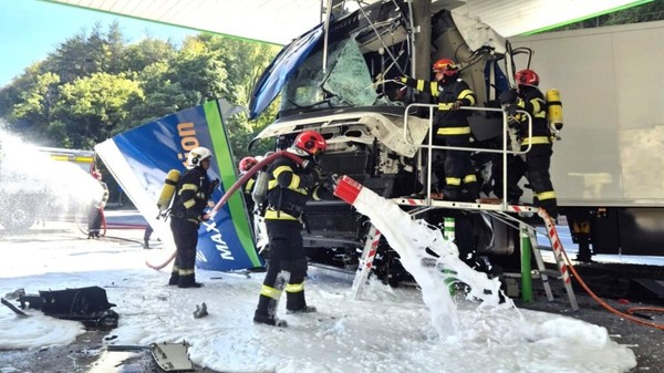Pompieri în echipament de protecție acționând cu spumă de stingere la locul accidentului în care cabina unui camion a fost distrusă în urma impactului cu structura unei benzinării.