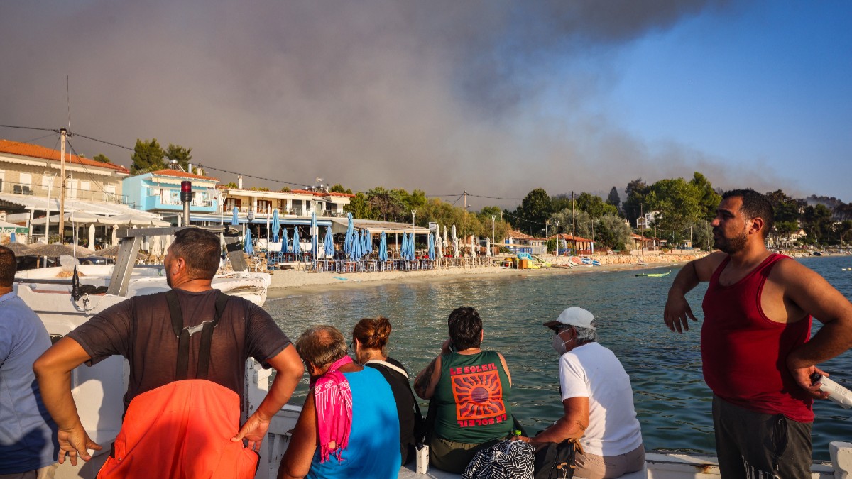 Imaginea prezintă un grup de persoane aflate pe o ambarcațiune pe apă, privind spre o zonă de coastă deasupra căreia se ridică un nor dens de fum întunecat provocat de un incendiu.
