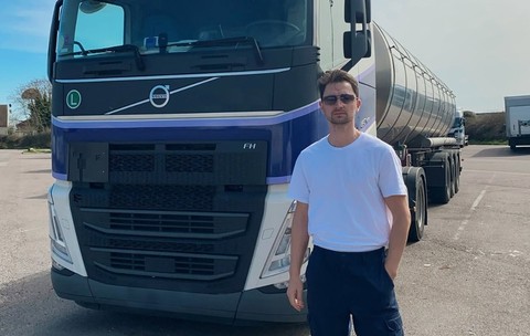 Un bărbat îmbrăcat în tricou alb, cu ochelari de soare, în fața unui camion cisternă Volvo într-o parcare.