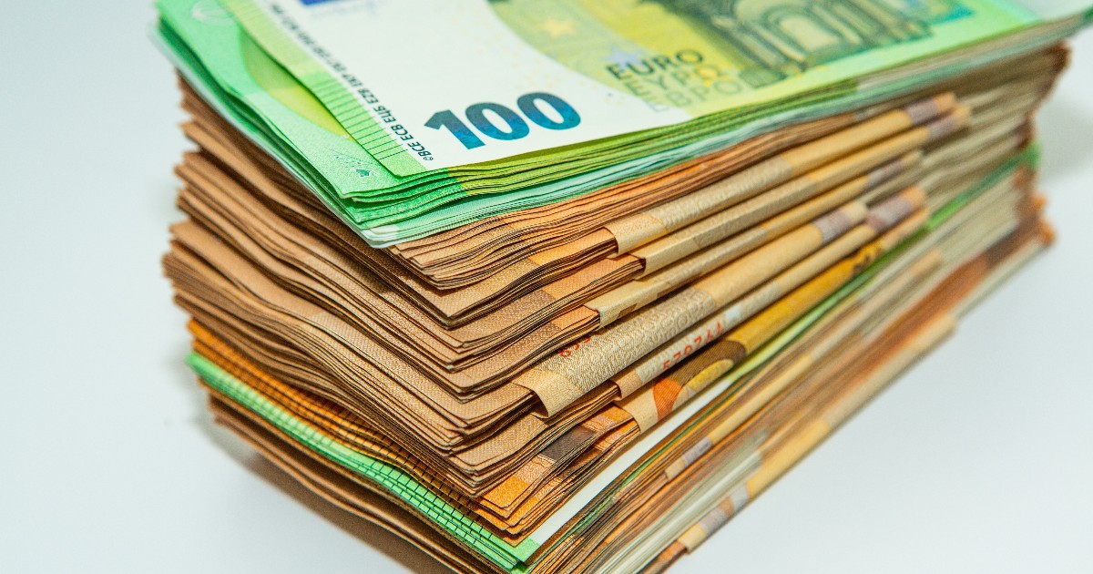 Imaginea prezintă un teanc gros de bancnote Euro, așezat pe o suprafață albă, indicând o sumă considerabilă de bani. Teancul este format preponderent din bancnote de 50 de Euro (cu nuanțe portocalii-maronii), cu una sau două bancnote de 100 de Euro (verzi) vizibile deasupra.