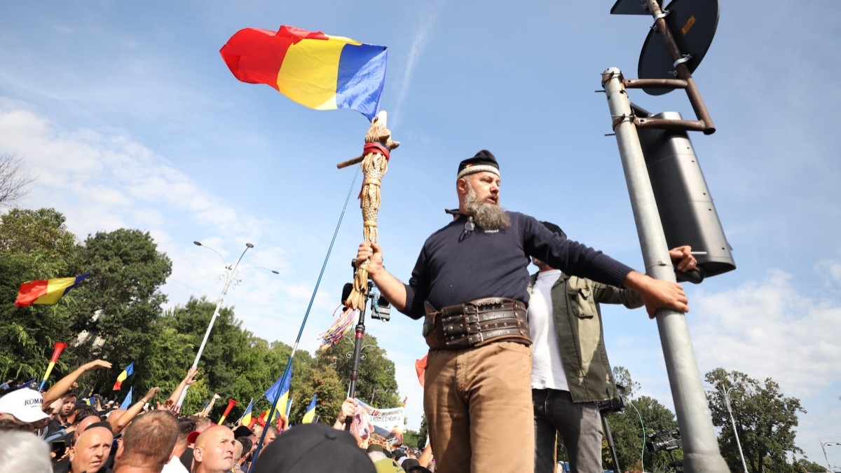 Un bărbat cu barbă și brâu din piele, purtând un toiag decorat și un steag al României, urcat la înălțime deasupra unei mulțimi de protestatari.