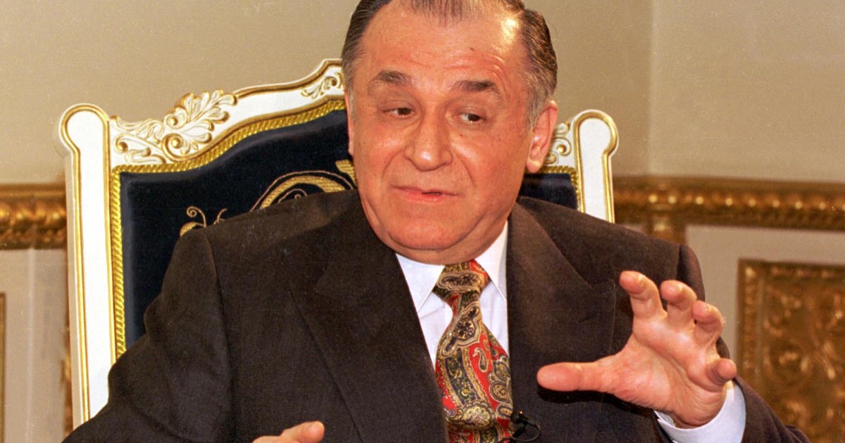 Imagine cu Ion Iliescu în timp ce stă pe un scaun. Fostul președinte a fost surprins cu mâinile ridicate. Poartă un costum negru și o cămașă albă
