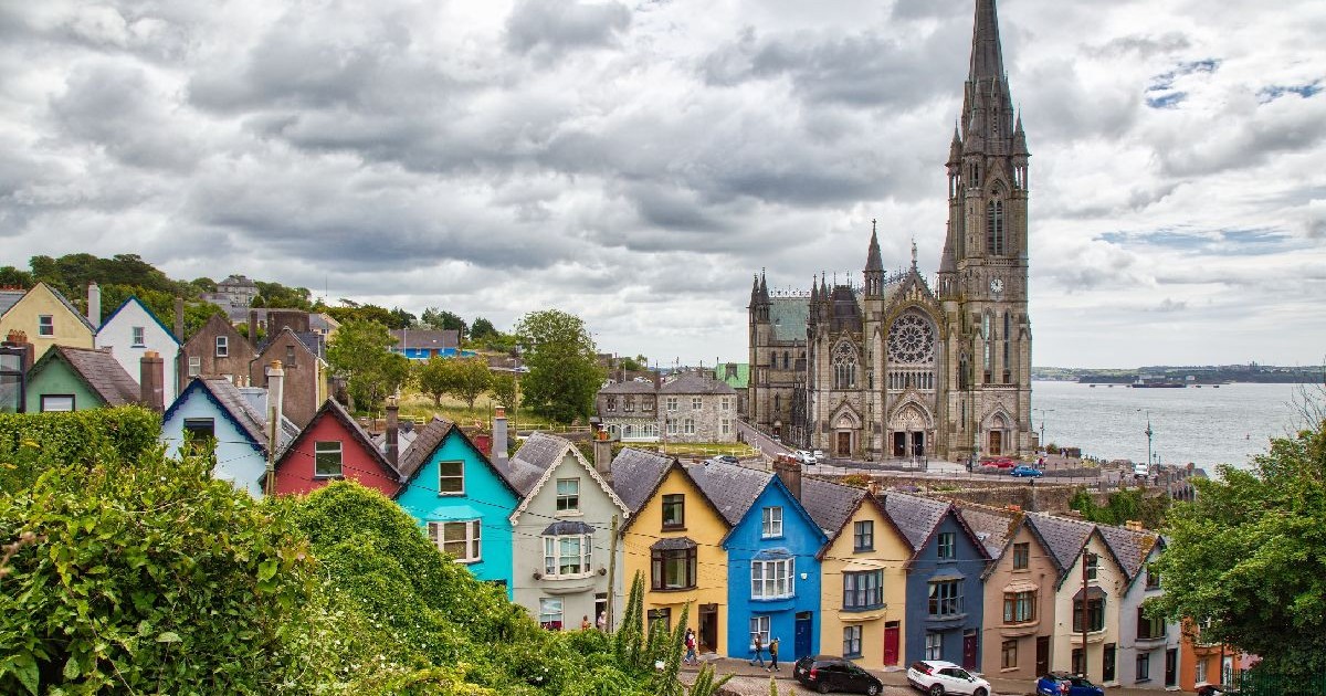 Imaginea prezintă un rând pitoresc de case viu colorate, cunoscute sub numele de „Deck of Cards”, situate în Cobh, Irlanda, având pe fundal impunătoarea Catedrală Sfântul Colman și apele portului sub un cer noros.