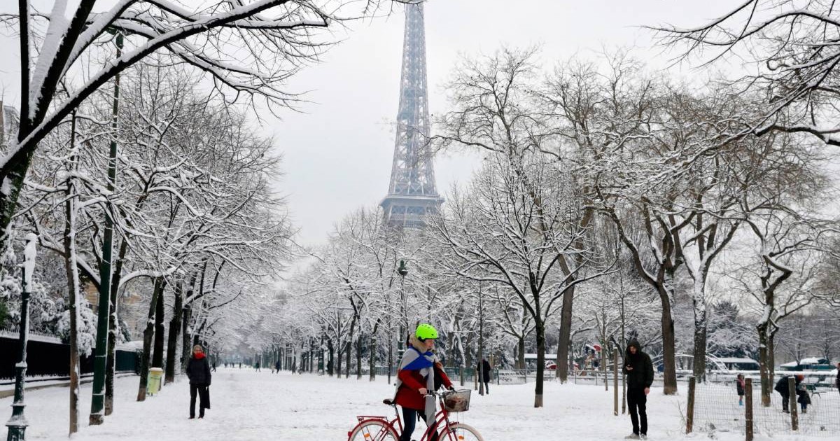 Parc in centrul Parisului, se vad strazi si copaci acoperiti de zapada, in fundal Turnul Eiffel, oameni se plimba, un tanar pe bicicleta cu o casca verde in centrul imaginii.