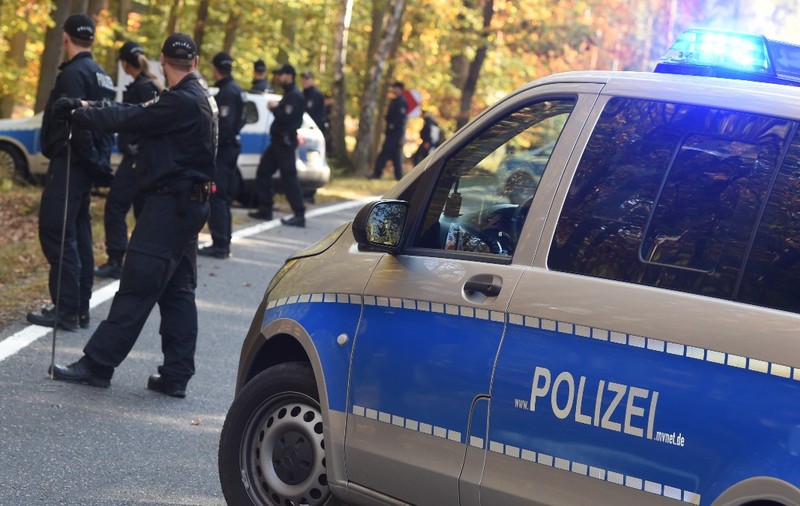 Mai mulți polițiști germani în uniforme desfășurând o operațiune de căutare sau securizare pe un drum de lângă o pădure, având o autospecială de poliție cu girofarul aprins în prim-plan.