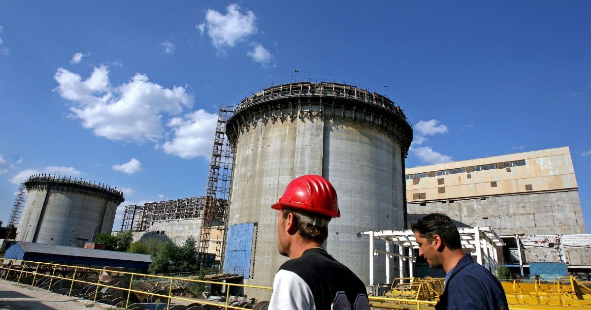 Centrala Nucleară de la Cernavodă - Doi muncitori văzuți din profil îmbrăcați în tricouri - unul alb cu negru și unul negru - trec prin fața unui Reactor. Muncitorul cel mai apropiat de cameră poartă o cască de siguranță roșie