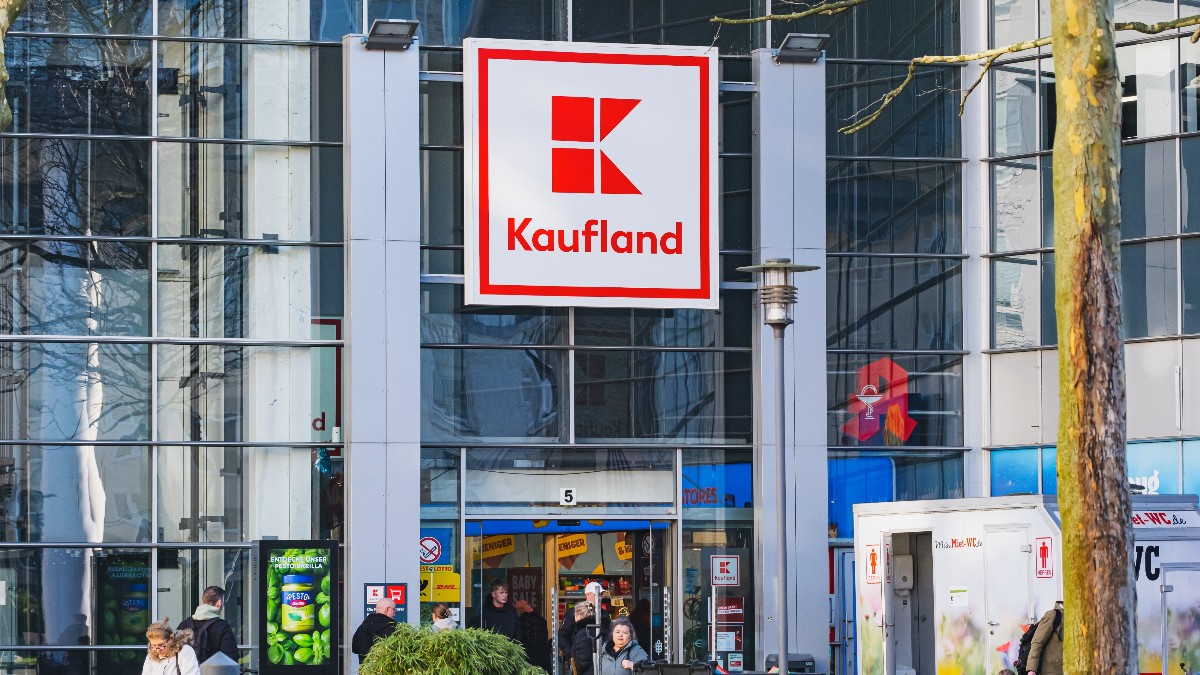 Imaginea prezintă intrarea unui magazin Kaufland, cu sigla roșie distinctivă afișată pe fațada din sticlă a clădirii. În fața magazinului se văd mai mulți clienți care intră sau ies, unii cu cărucioare de cumpărături, într-un cadru urban tipic, ce sugerează activitatea zilnică intensă a supermarketului.
