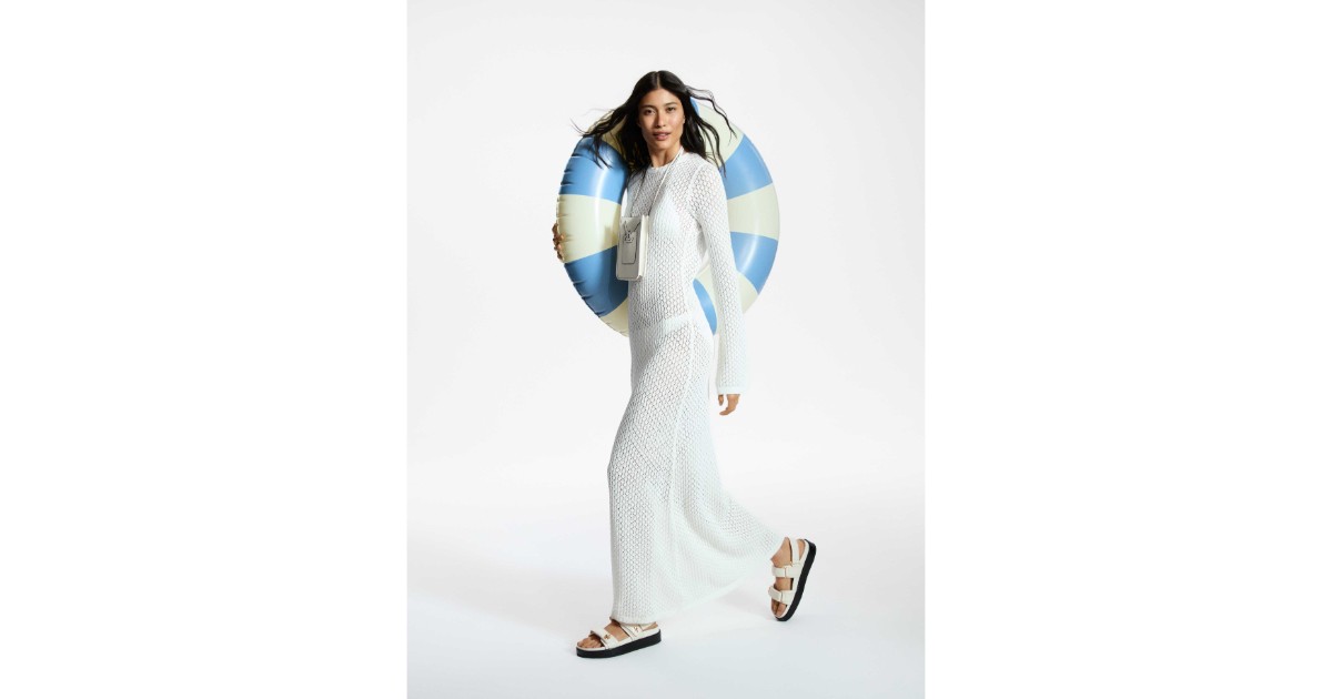 Colecție MODIVO Summer 2025. Model, fata cu parul lung si brunet, imbracata intr-un caftan de plasa alb, lung, cu manecu lungi, tine pe umar un colac de piscina mare, alb, cu dulgi albastru deschis