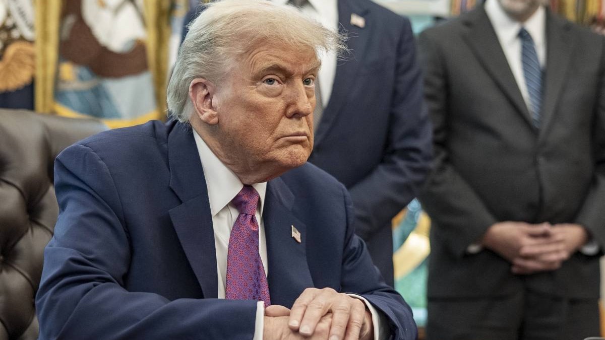 Președintele american Donald Trump în timp ce stă la biroul său de la Casa Albă, are mâinile încrucișate pe birou, poartă un costum albastru și o cravată mov.
