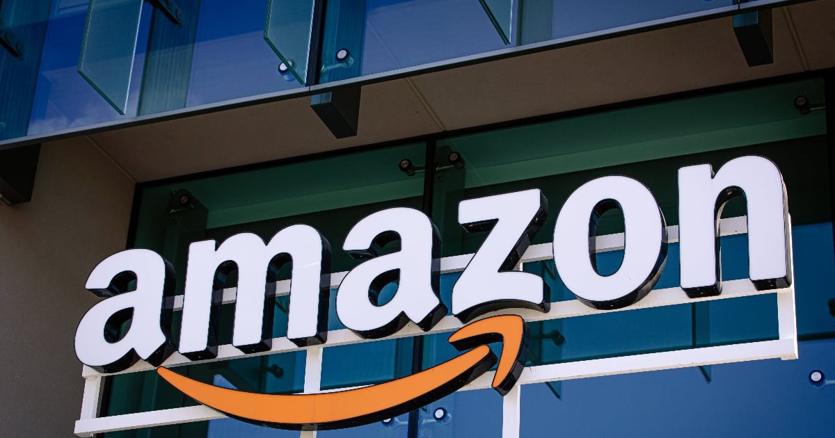 Imaginea surprinde un logo Amazon tridimensional montat pe exteriorul unei clădiri de birouri cu fațadă din sticlă. Literele albe ale numelui companiei sunt așezate deasupra săgeții portocalii stilizate, care simbolizează un zâmbet și se întinde de la litera 'a' la 'z'.