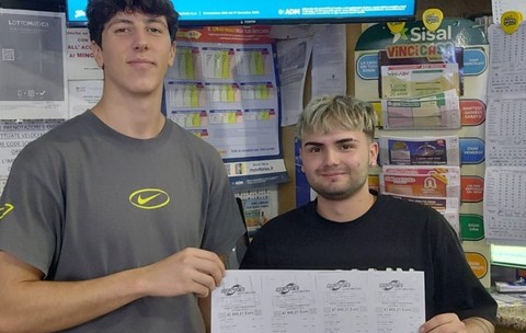 Doi tineri care zâmbesc și țin împreună mai multe bilete de pariu/loterie într-o agenție de jocuri de noroc, pe bilete fiind vizibil un câștig de 47.950,21 euro.