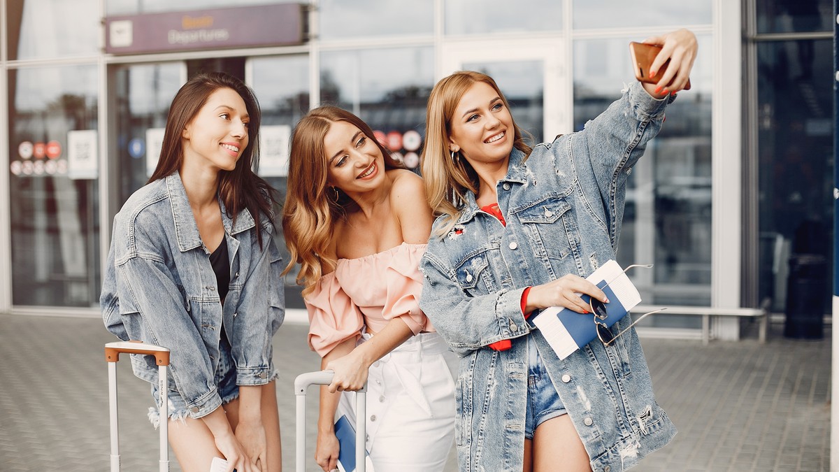 trei femei care stau apropiate în fața unei clădiri moderne cu ferestre și uși mari din sticlă, asemănătoare terminalului unui aeroport sau unei gări. Femeia din partea dreaptă ține un telefon mobil ridicat, făcând un selfie grupului, în timp ce toate trei zâmbesc și privesc spre obiectivul telefonului. Ele țin în mâini pașapoarte și documente de călătorie, având alături bagaje cu roți și mânere extensibile, ceea ce indică faptul că se pregătesc de o plecare. Sunt îmbrăcate lejer, în ținute specifice sezonului cald, incluzând jachete din denim, pantaloni scurți, o bluză cu umerii goi și o fustă. Suprafața pe care se află este pavată cu dale dreptunghiulare.