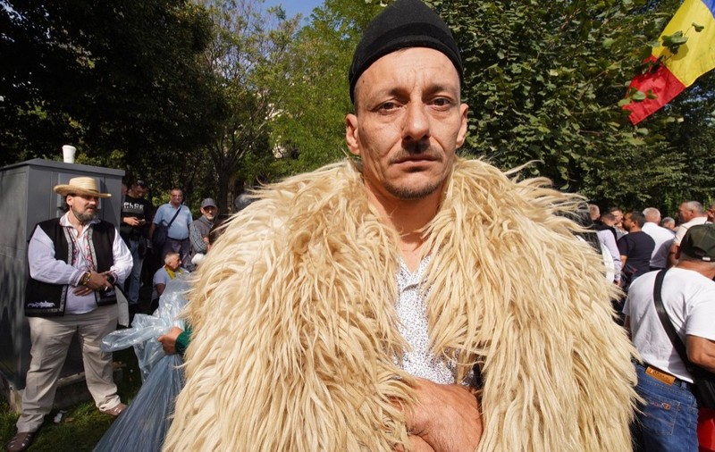 Un bărbat purtând o căciulă neagră și un cojoc din blană de oaie, privind fix spre cameră în mijlocul altor participanți la un protest în aer liber, unde se observă și un steag al României.