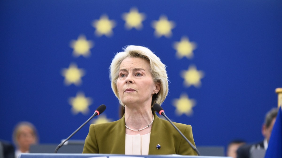 Imaginea o arată pe Ursula von der Leyen vorbind la un pupitru. Are părul scurt, blond și poartă un sacou verde-măsliniu peste un top de culoare crem. Un colier de perle este vizibil la gât. Fundalul este un steag albastru cu un cerc de stele galbene. Două microfoane se află în fața ei Foto: Profimedia
