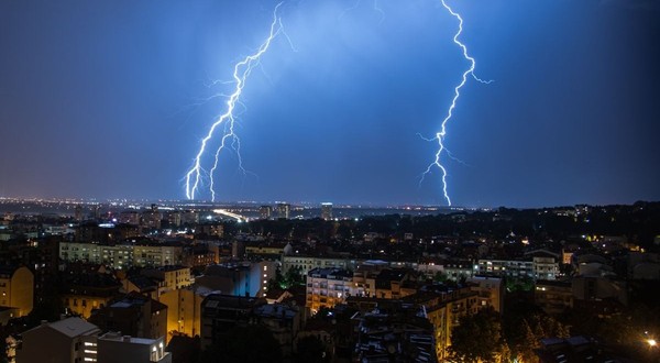 Furtună cu fulgere și cer înnorat peste un oraș, cu două fulgere care lovesc pământul în fundal